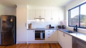 A modern kitchen with white cabinets, a built-in oven, a stainless steel refrigerator, and a large window providing natural light. - 428 Eastbourne Road · Rosebud Garden Cottage (Capel Sound)