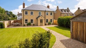 A two-story house with a well-maintained lawn, a wooden fence, and a garage. - Essitone: Cinema Room, Games Room, Gym & More! (Ashton Keynes)