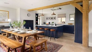 A modern kitchen with a central island, wooden beams, and a dining area with a table set for six. - Essitone: Cinema Room, Games Room, Gym & More! (Ashton Keynes)