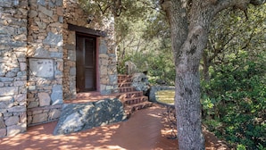 A stone building with a wooden door, steps leading up to it, and a paved area with a tree in the foreground.