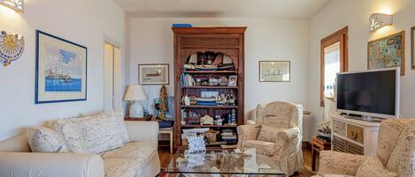 A living room with a glass coffee table, two patterned armchairs, a wooden bookshelf, and a television.