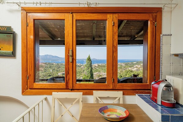 Una habitación con ventana de madera que da a un paisaje pintoresco, una mesa con una cesta tejida de colores y una cafetera roja en la encimera.