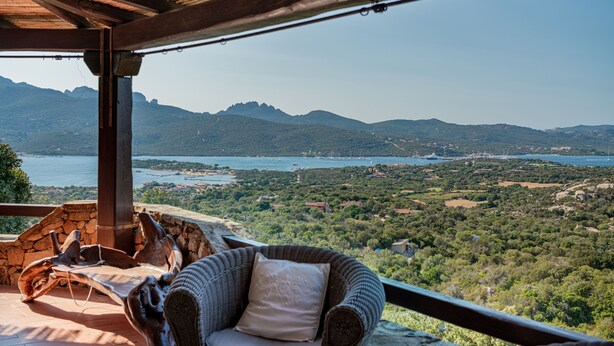A wicker lounge chair with a cushion, a stone wall, and a view of a mountainous landscape with a body of water in the distance.