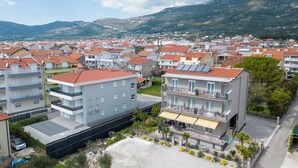 A multi-story hotel with balconies, a parking area, and a view of a residential area with red-roofed houses and mountains in the background. - Villa Vinovo 1 Bedroom Apartment (Kastela)