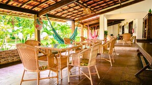 A covered outdoor seating area with wicker chairs and tables, a hammock, and a view of lush greenery. - Pousada da Mércia (Salvador)