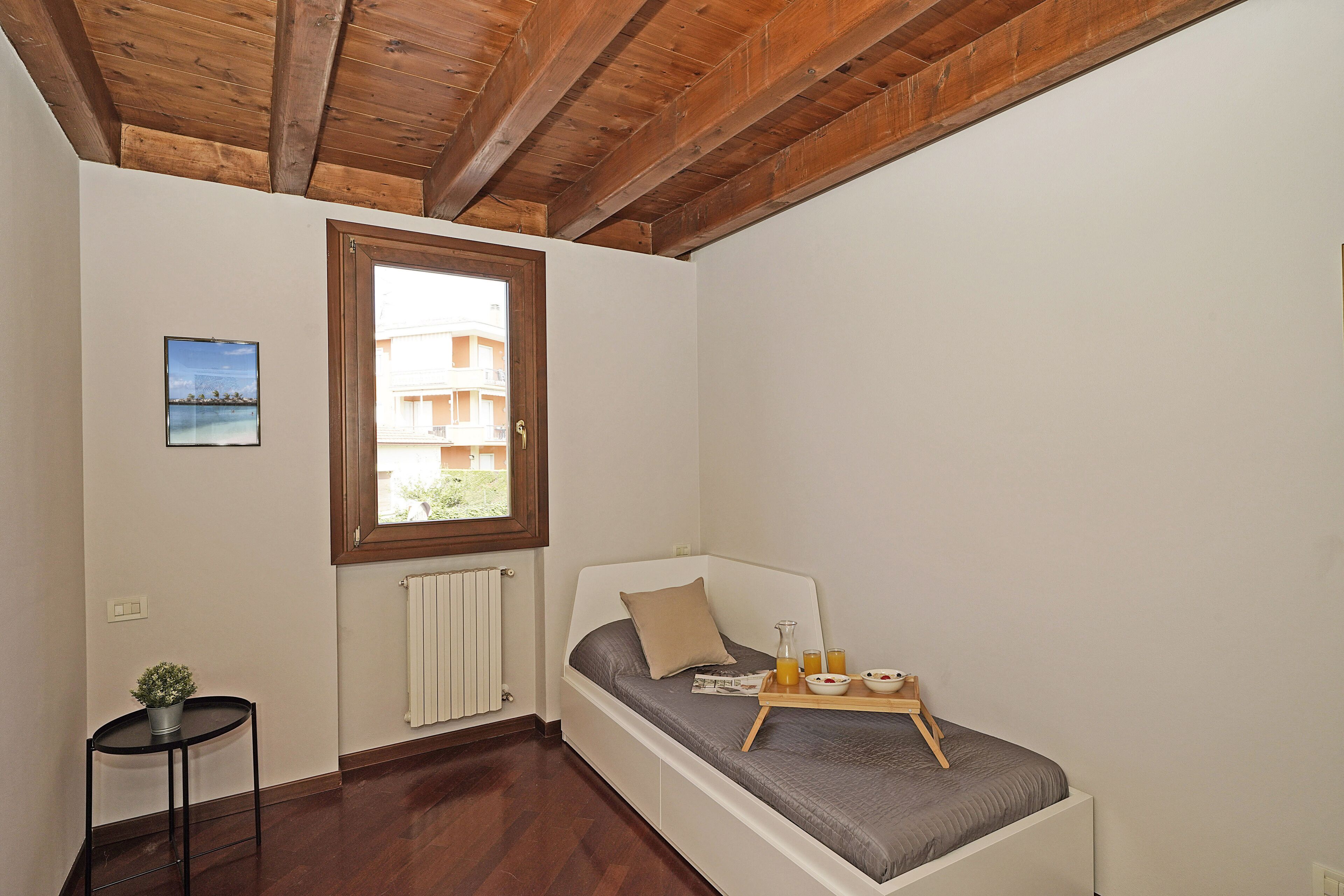 A small, single-bed room with a wooden ceiling, a window with a view of buildings, a small table with a tray of breakfast items, and a potted plant.