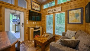 A cozy cabin interior with a wooden interior, a fireplace, a television, and a large window with blinds. - Grin & Bear It: brand new tiny home 1 bdr + loft w/ hot tub (Broken Bow)