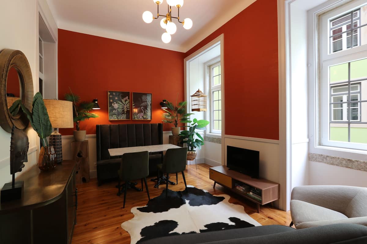 A modern living room with a red wall, wooden flooring, a cowhide rug, a dining area with a table and chairs, a television, and a large window with a view.
