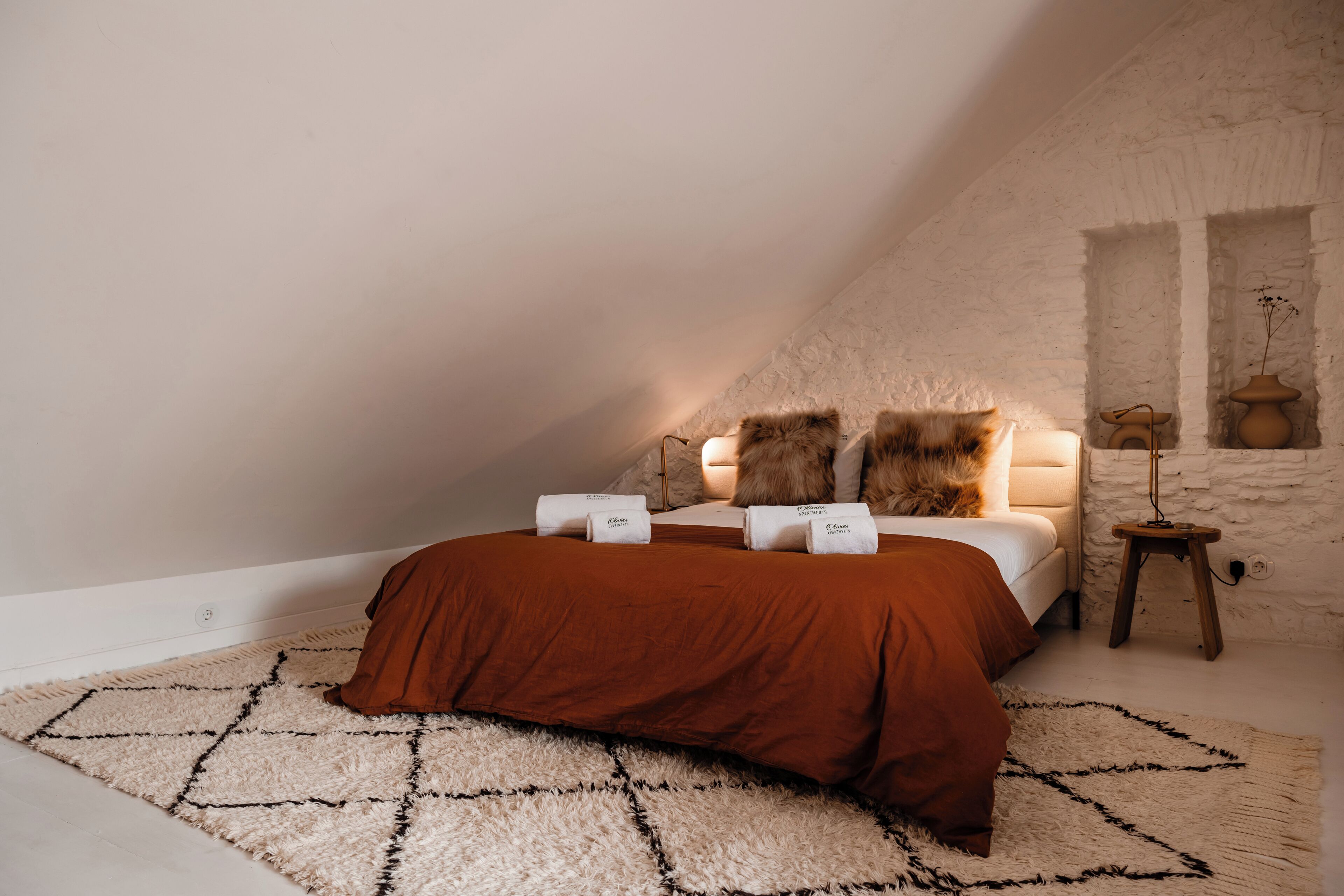 Ein Schlafzimmer mit schrägverdeckter Decke, einem Bett mit brauner Bettdecke, zwei Kissen, einem kleinen Holfnachtkästchen mit einer Lampe und einem weißen Teppich mit Struktur.