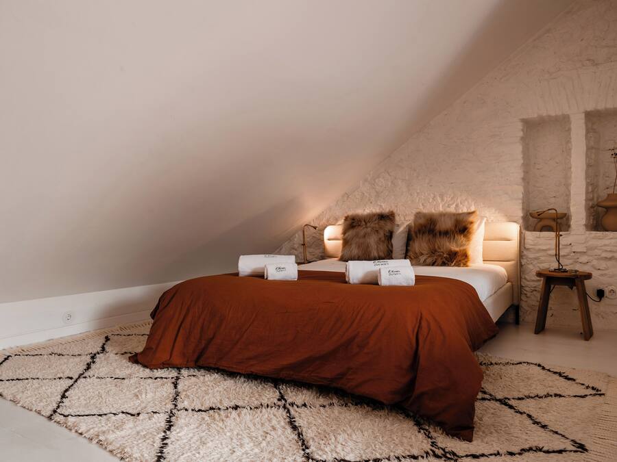 Ein Schlafzimmer mit schrägverdeckter Decke, einem Bett mit brauner Bettdecke, zwei Kissen, einem kleinen Holfnachtkästchen mit einer Lampe und einem weißen Teppich mit Struktur.