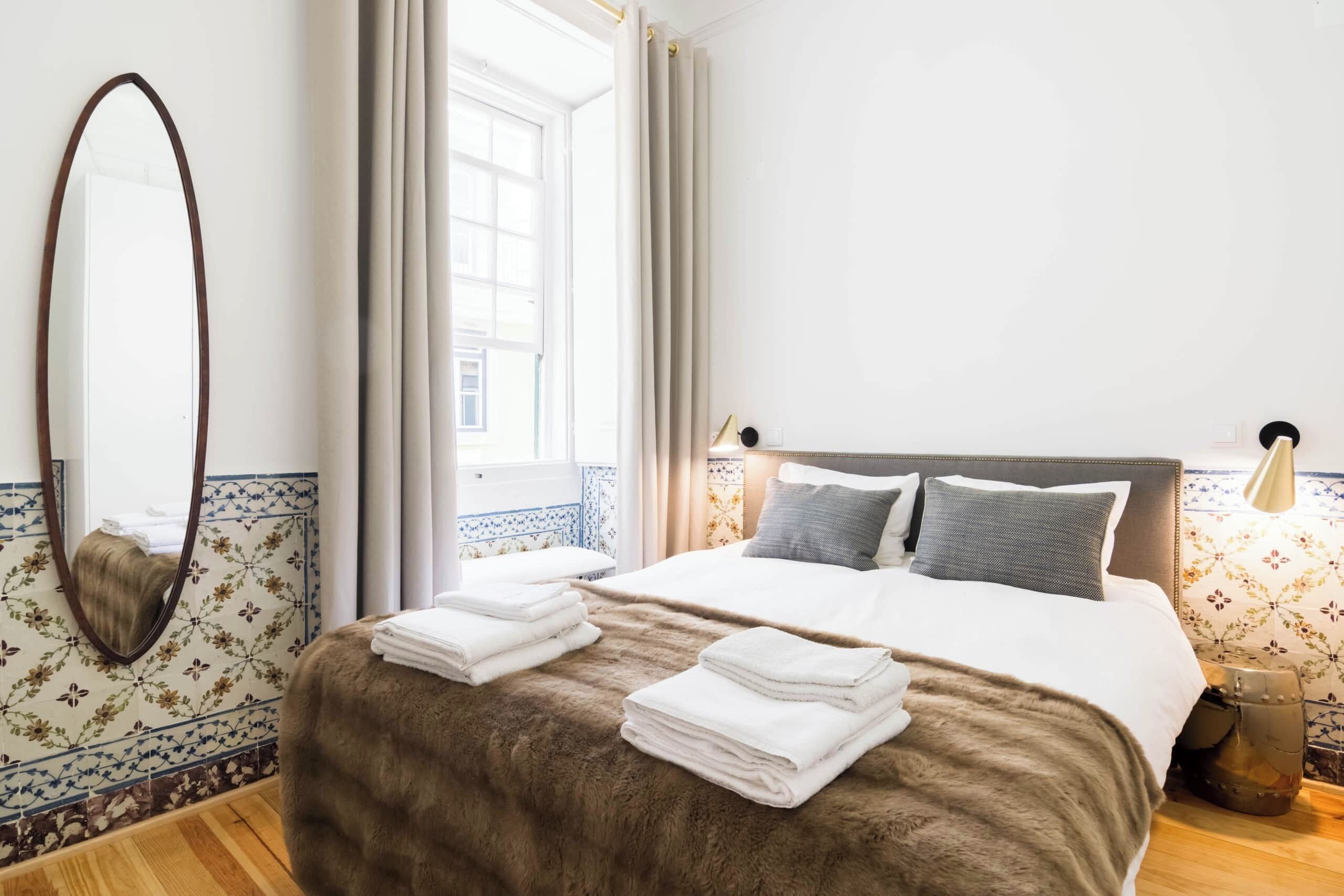 Una camera d'albergo con un letto grande, uno specchio, una finestra con tende e un comodino con una lampada.