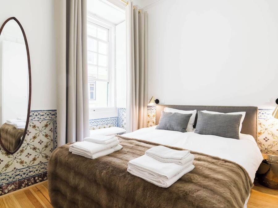 Una camera d'albergo con un letto grande, uno specchio, una finestra con tende e un comodino con una lampada.