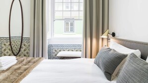 Hotellihuone, jossa on sänky, yöpöytälamppu, ikkuna, josta on näkymä rakennukseen, ja peili seinällä.