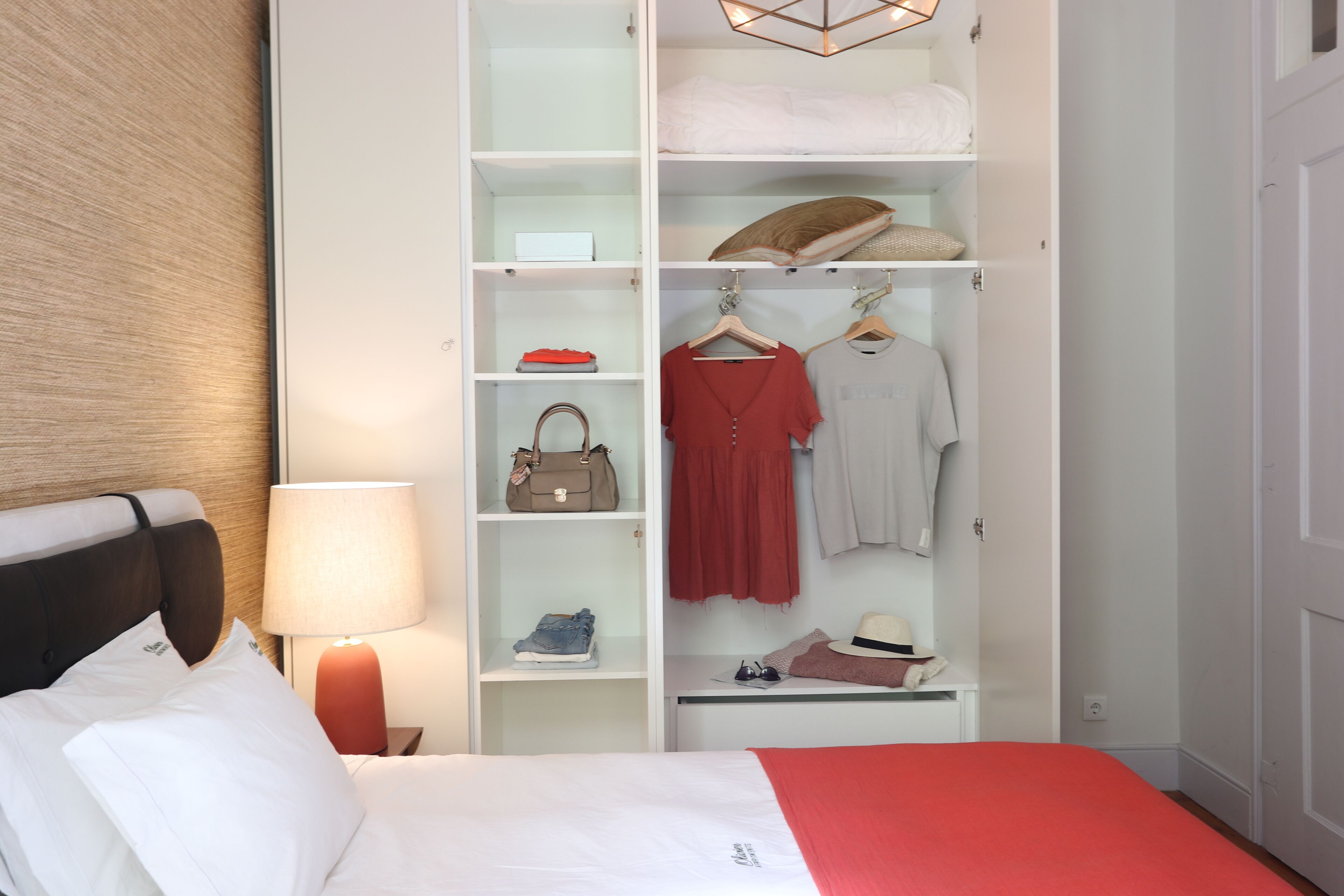 A hotel room with a neatly organized wardrobe, a bed with white bedding, and a bedside lamp.