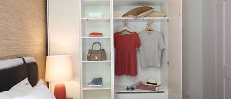 A hotel room with a neatly organized wardrobe, a bed with white bedding, and a bedside lamp.