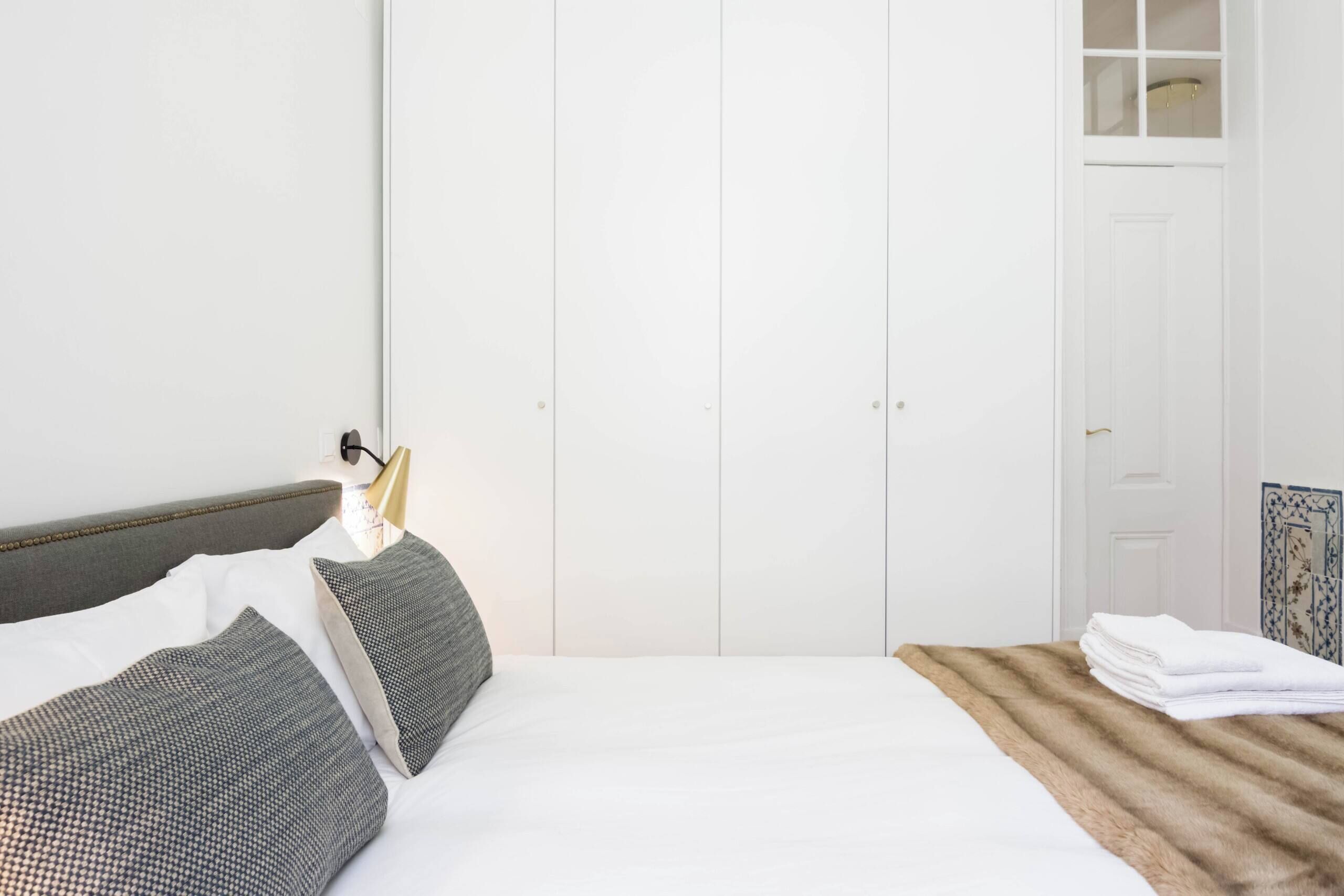 A neatly made bed with white linens and grey patterned pillows, a wooden headboard, and a white wardrobe with a small window.