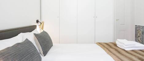 A neatly made bed with white linens and grey patterned pillows, a wooden headboard, and a white wardrobe with a small window.