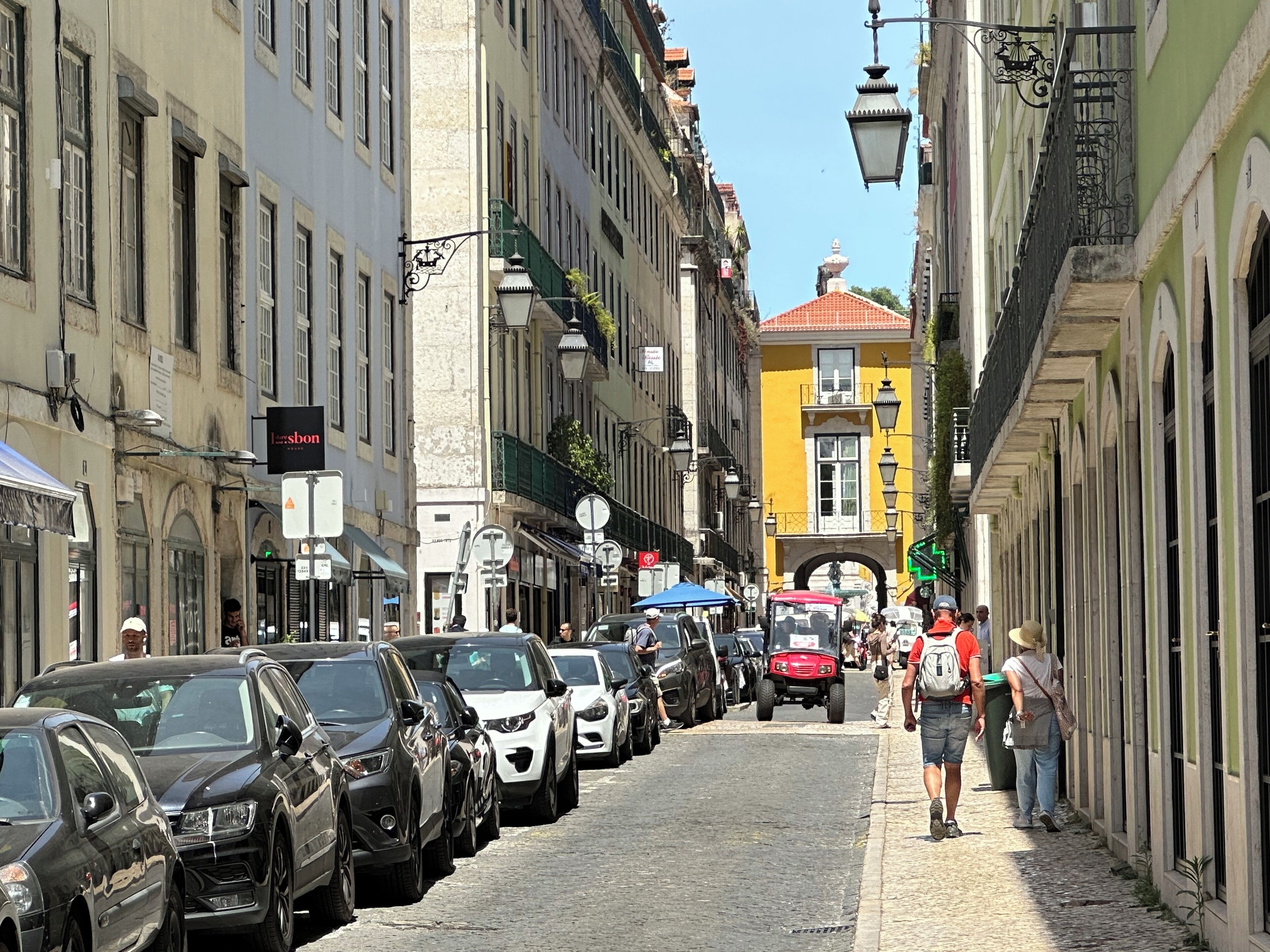 Une rue étroite pavée, bordée de voitures garées et de piétons.