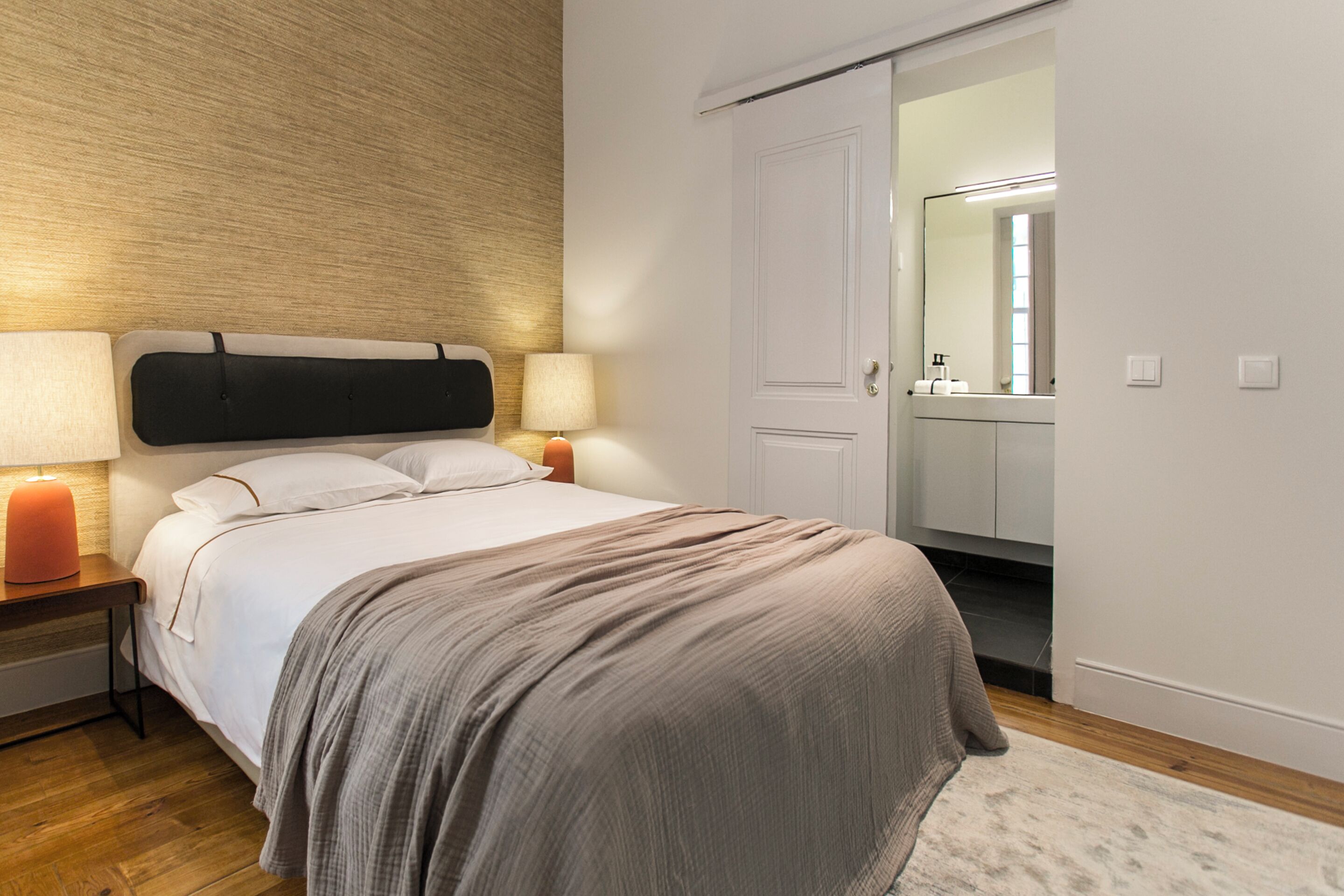 Quarto bem arrumado com cama, abajures, mesa de cabeceira, cabeceira de madeira e porta que dá para um banheiro.