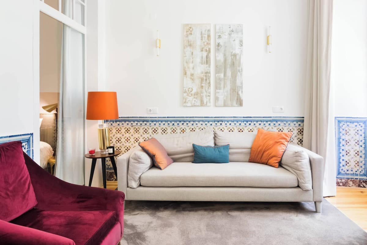 A living room with a sofa, a red chair, a small table with a lamp, and two wall-mounted light fixtures.