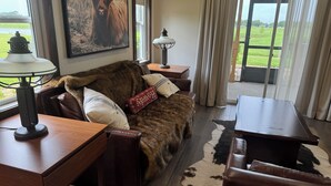 A cozy living room with a fur-covered sofa, a wooden coffee table, and a lamp. A framed picture of a yak is on the wall, and a window offers a view of a green landscape. - Cozy cabin with 2 bedrooms and WiFi, AC in soothing River Ranch (River ranch)