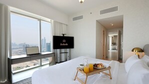 Habitación de hotel con cama, escritorio con una laptop, televisor, bandeja de desayuno y vista a la ciudad.