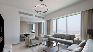 Una sala de estar moderna con un sofá, sillones, una mesa de centro y un comedor con una mesa y sillas. Amplias ventanas ofrecen vistas de la ciudad.
