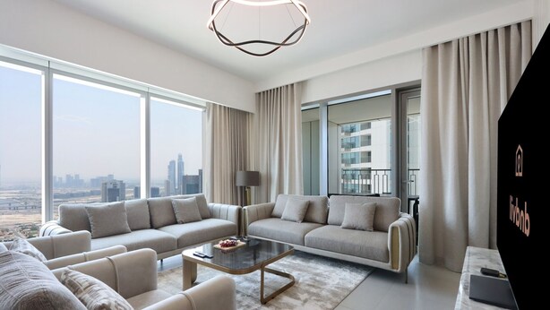 Una sala de estar moderna con un sofá, una mesa de centro y vistas a un paisaje urbano a través de amplios ventanales.
