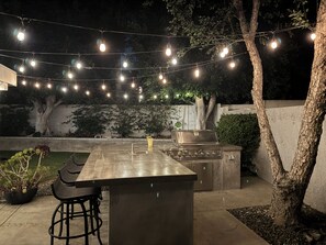 Een terras buiten met een bar, krukken, een barbecue en lichtjes.