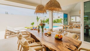 A spacious dining area with a long wooden table, wicker chairs, and a large window overlooking a swimming pool. - Luxury Residence Palm Beach | Villa Amandari (Noord)