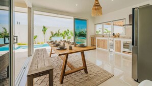 A modern kitchen with wooden cabinets, a stainless steel refrigerator, and a large window overlooking a pool area. - Luxury Residence Palm Beach | Villa Amandari (Noord)