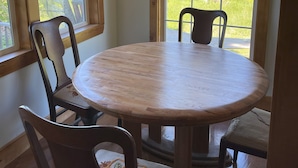 A room with a round wooden table and four chairs, large windows with a view of greenery, and wooden ceiling beams.