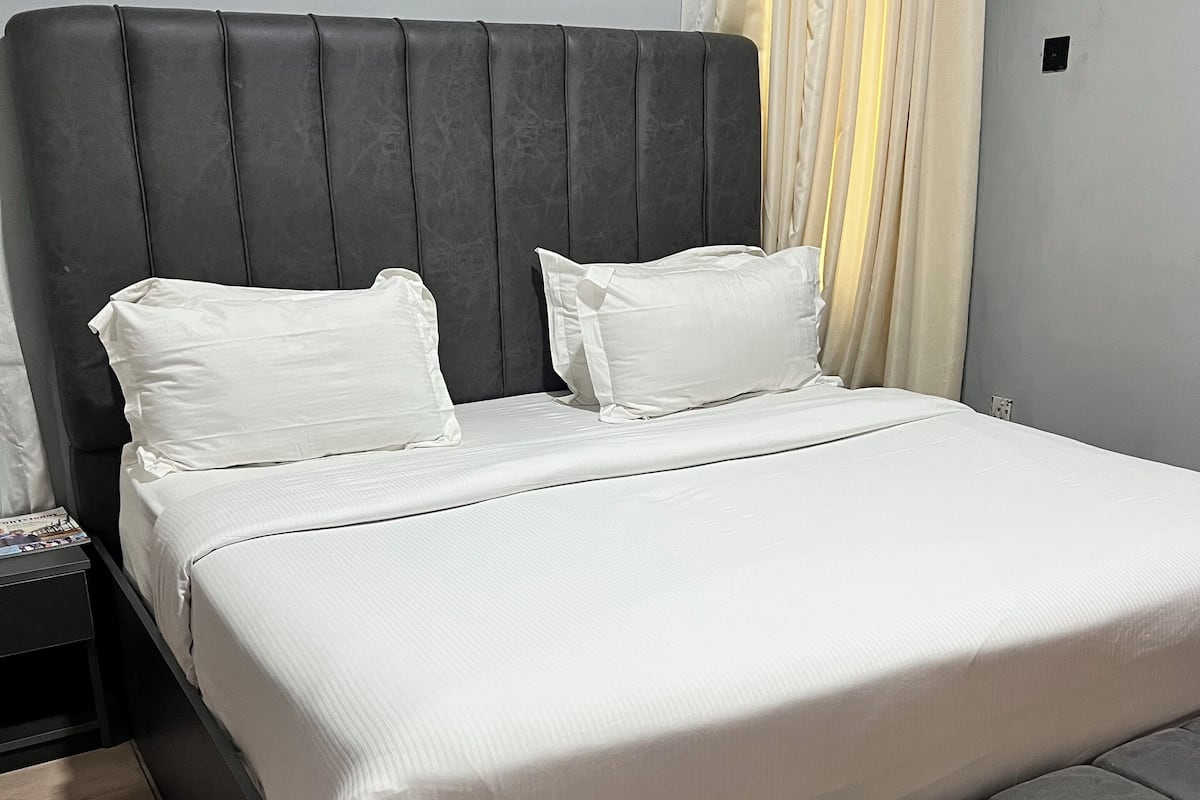 Un lit avec une tête de lit et un pied de lit recouverts d’un tissu gris, et une literie blanche.