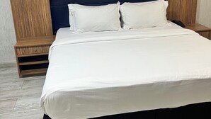 A modern bedroom with a large bed, dark headboard, wooden nightstands, and a light-colored floor.