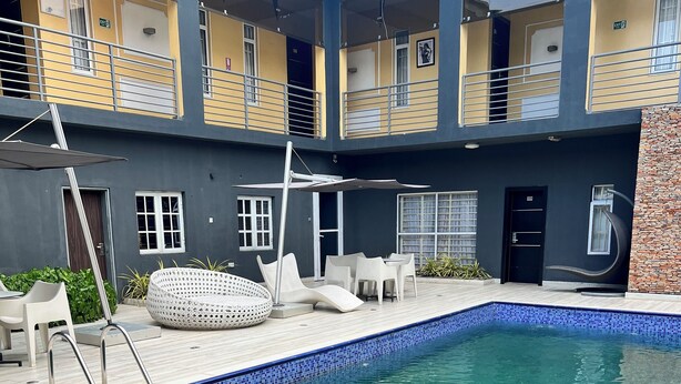 A modern indoor swimming pool with clear blue water, surrounded by a wooden deck and furnished with white chairs and a lounge area.