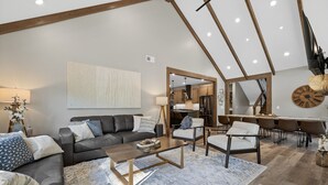 Un salón espacioso con un sofá de cuero oscuro, una mesa de centro de madera y una alfombra grande. Se ve una cocina a través de un arco abierto, y en el fondo hay un comedor con mesa y sillas de madera.