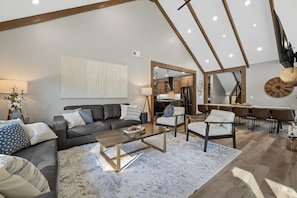 Een ruime woonkamer met een donker leren bank, een houten salontafel en een groot vloerkleed. Een keuken is zichtbaar door een open doorgang, en in de achtergrond bevindt zich een eethoek met een houten tafel en stoelen.