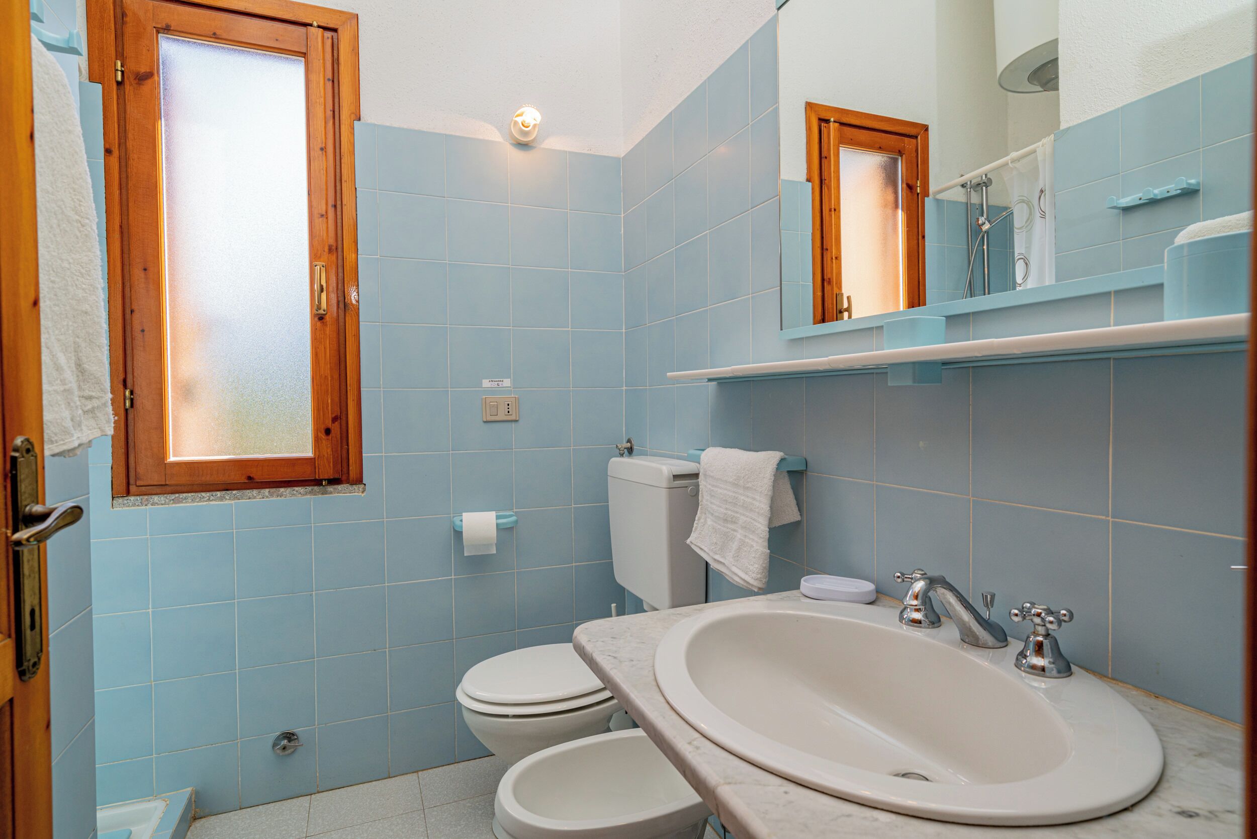 Ein Badezimmer mit blauen Fliesen, einem Fenster mit Holzrahmen, einem weißen Waschbecken und einer Toilette.