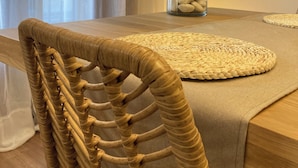 Una mesa de comedor con un mantel tejido, un florero transparente con una planta y una silla con respaldo de mimbre.
