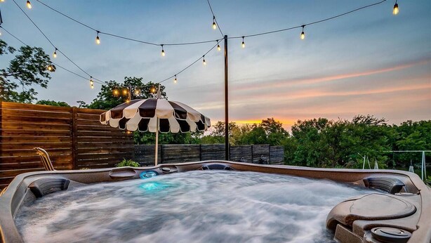 Un bain à remous avec vue sur le coucher de soleil, entouré d’une clôture en bois et illuminé de guirlandes lumineuses.