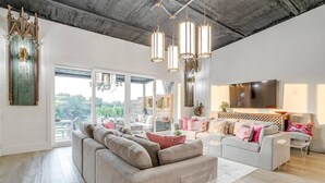 Un salon spacieux avec un grand canapé, un petit divan et un téléviseur. On y trouve plusieurs luminaires suspendus et une grande fenêtre qui offre une vue sur l’extérieur.