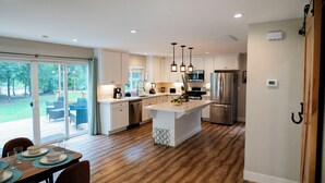 A modern kitchen with white cabinetry, a central island, and a dining area with a glass door leading to a patio. - Charming Spacious Riverside 2-bedroom house in welcoming Reno with AC, WiFi (Reno)