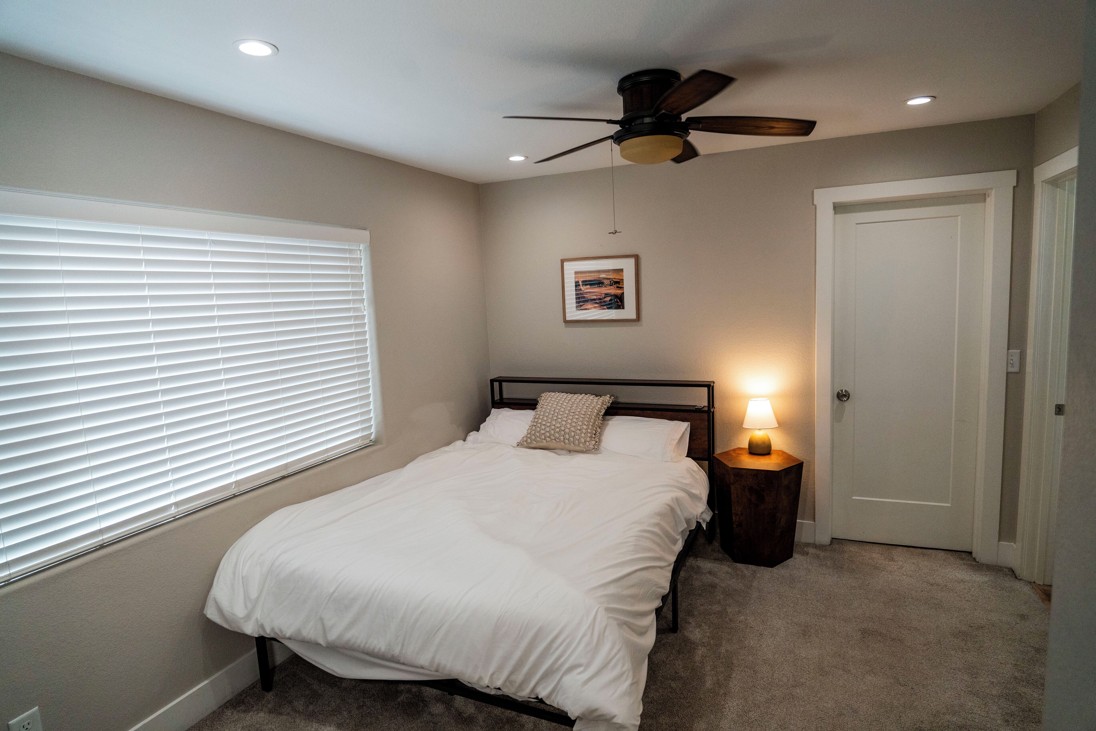 Un dormitorio con cama, ventilador de techo, mesita de noche con lámpara, una puerta y ventana con persianas.