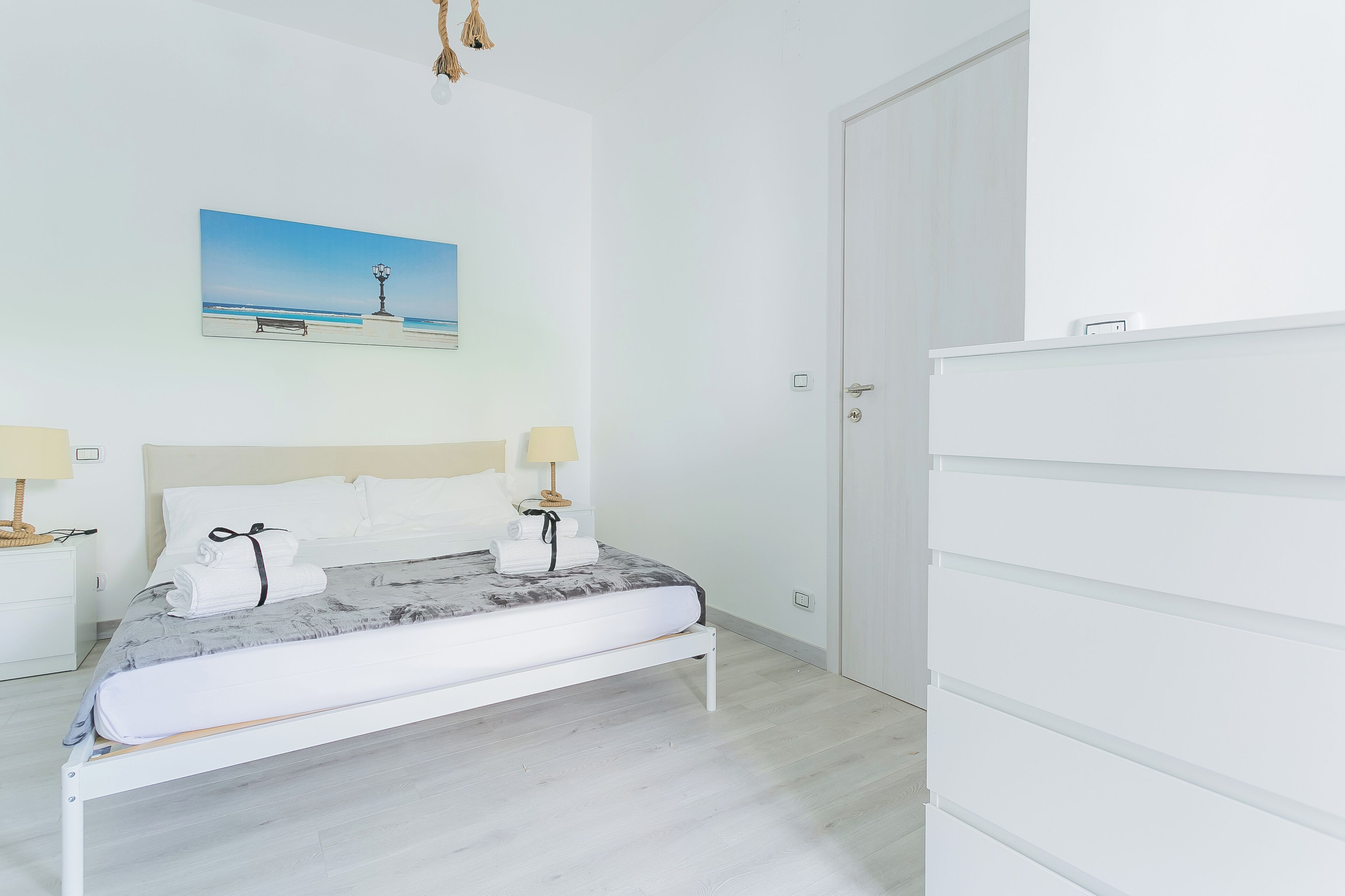 Une chambre à coucher bien rangée, avec un lit, des lampes de chevet, un tableau représentant une scène de plage et une armoire blanche.