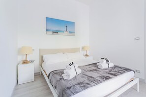 Une chambre d’hôtel avec un lit, deux tables de chevet avec des lampes, un tableau représentant une scène de plage et un banc.