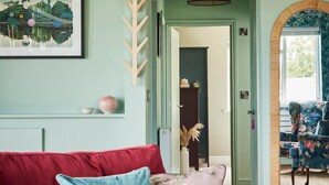 Un canapé rouge agrémenté de coussins bleus et roses, un tableau encadré, un porte-manteau en bois, un luminaire suspendu et, à travers une ouverture, on aperçoit une autre pièce avec une chaise bleue.