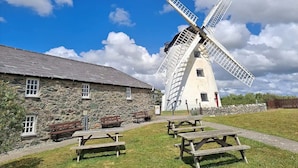 Un moulin à vent traditionnel, avec un bâtiment en pierre à côté, des tables de pique-nique et des bancs.