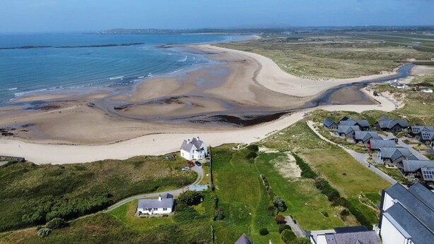 Vue aérienne d’une zone côtière avec une plage de sable, un champ herbeux et plusieurs maisons aux toits inclinés.