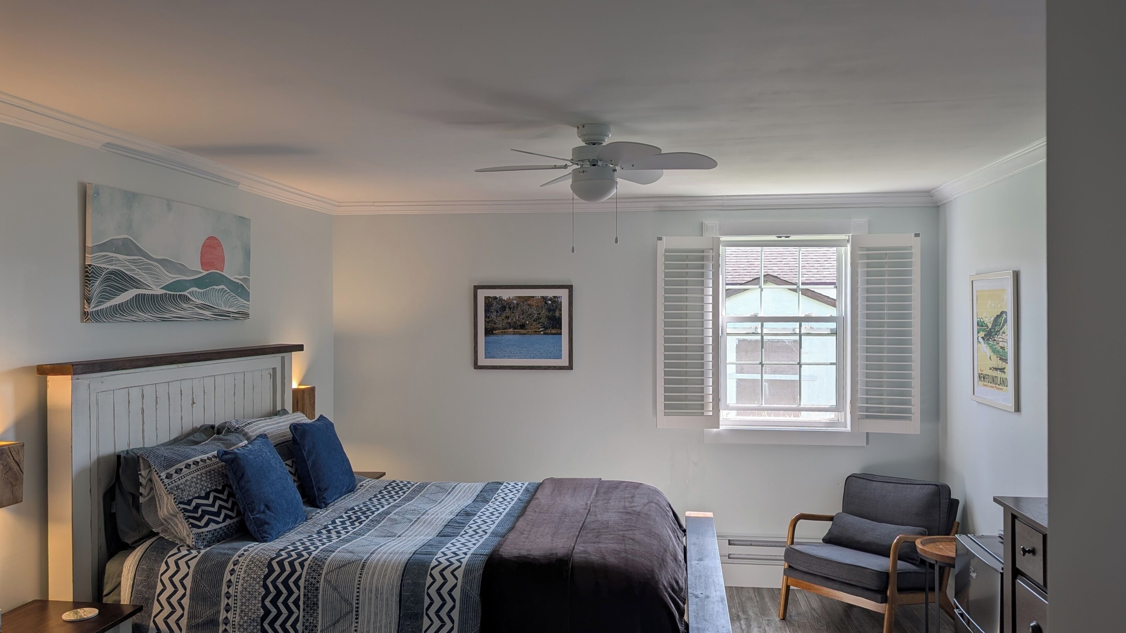 A bedroom with a bed, a chair, a window with shutters, a ceiling fan, and artwork on the walls.