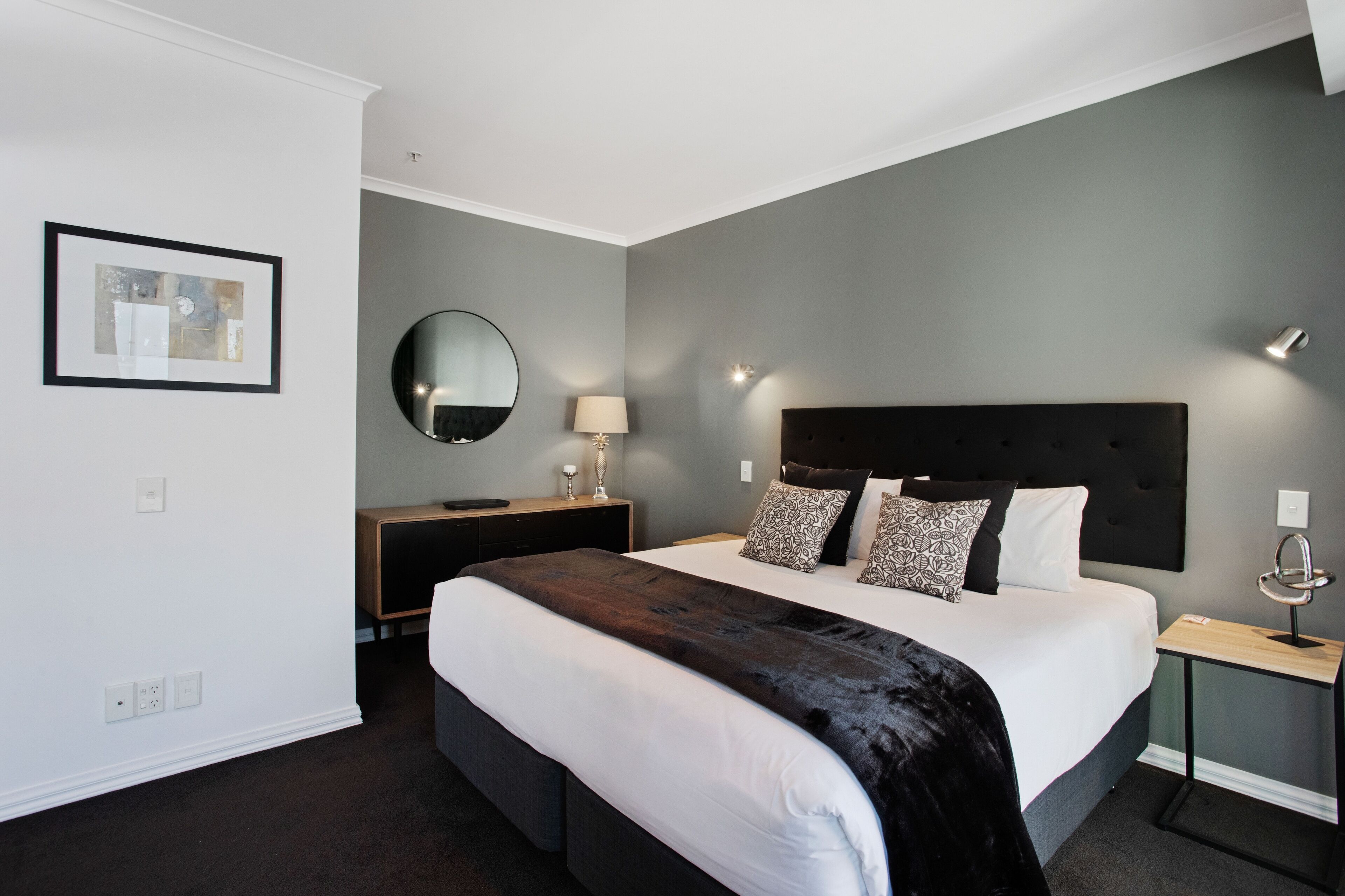 A modern hotel room with a large bed, a bedside table, a mirror, and a framed picture on the wall.